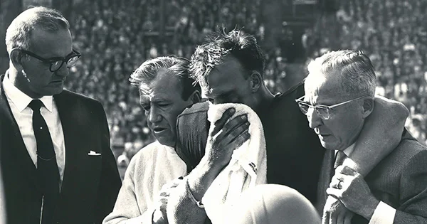 Black and white photo showing a football player being consoled by coaches and staff on the sidelines. Large crowd visible in the background.