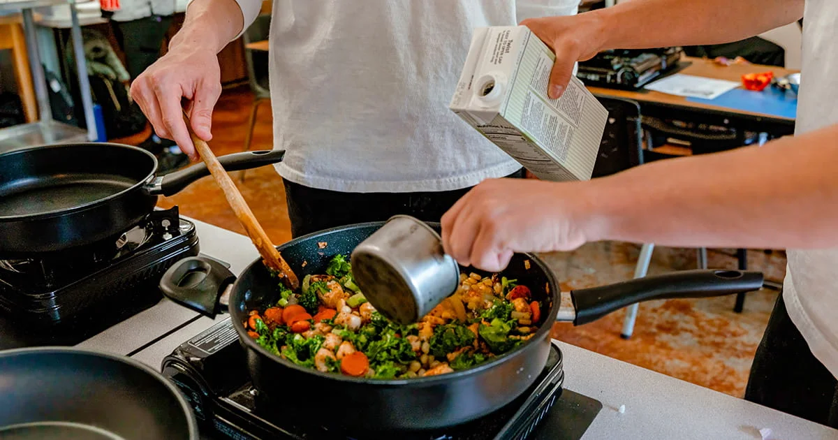 Two people cooking vegetables in a pan on a portable stovetop, one pouring liquid from a carton.