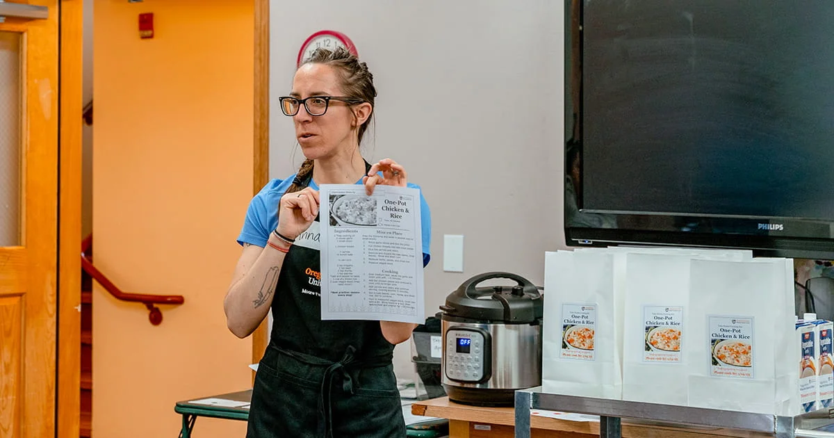Woman holding a recipe for "One-Pot Chicken & Rice" next to an Instant Pot and prepared meal kits. She's wearing glasses and an apron in a classroom setting.