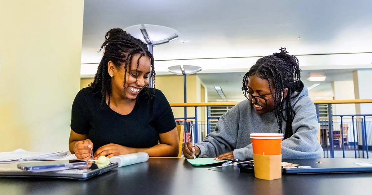 Students collaborate on a project in a library or study hall, working together and smiling. Represents teamwork and academic engagement.