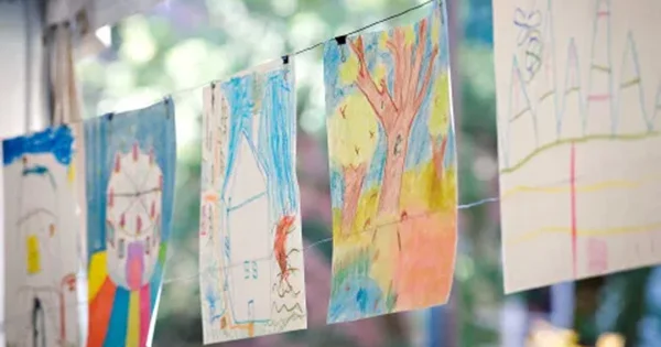 Children's crayon drawings of a tree, house, and Ferris wheel clipped to a string against a soft, blurred green background.