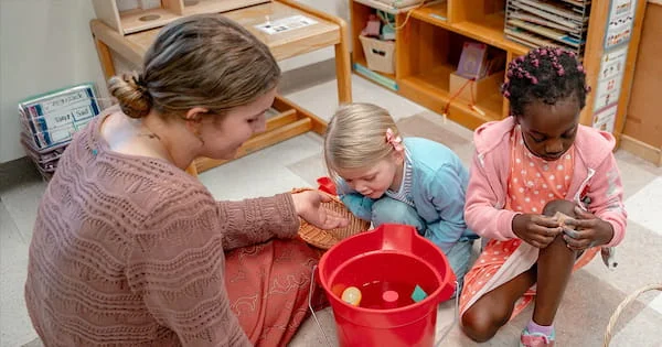 Preschool students engage in sensory play with a red bucket of water while a teacher assists them in a well-equipped classroom environment.