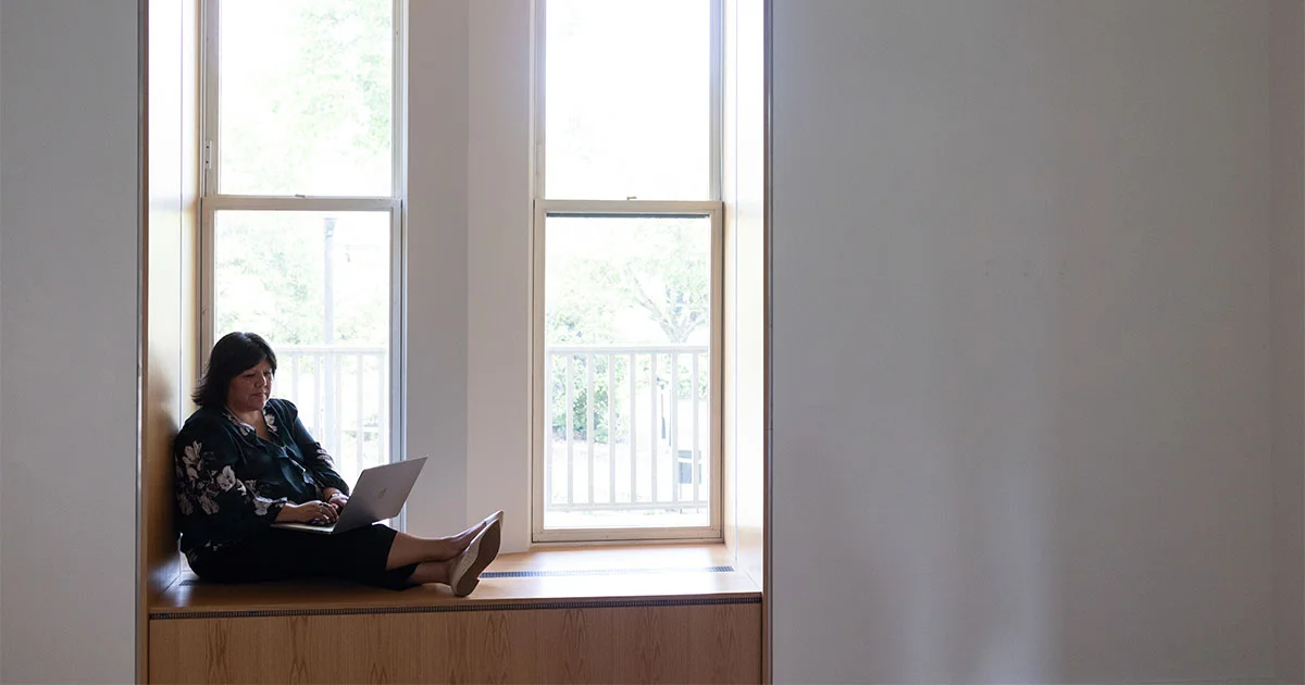 A woman sits in a window nook using a laptop, silhouetted by bright daylight from two large windows.