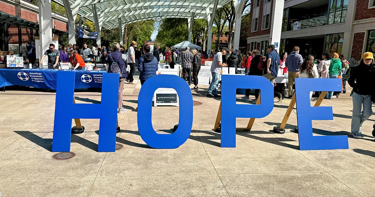 Large blue block letters spelling HOPE stand in a sunny outdoor plaza filled with people and event tables.