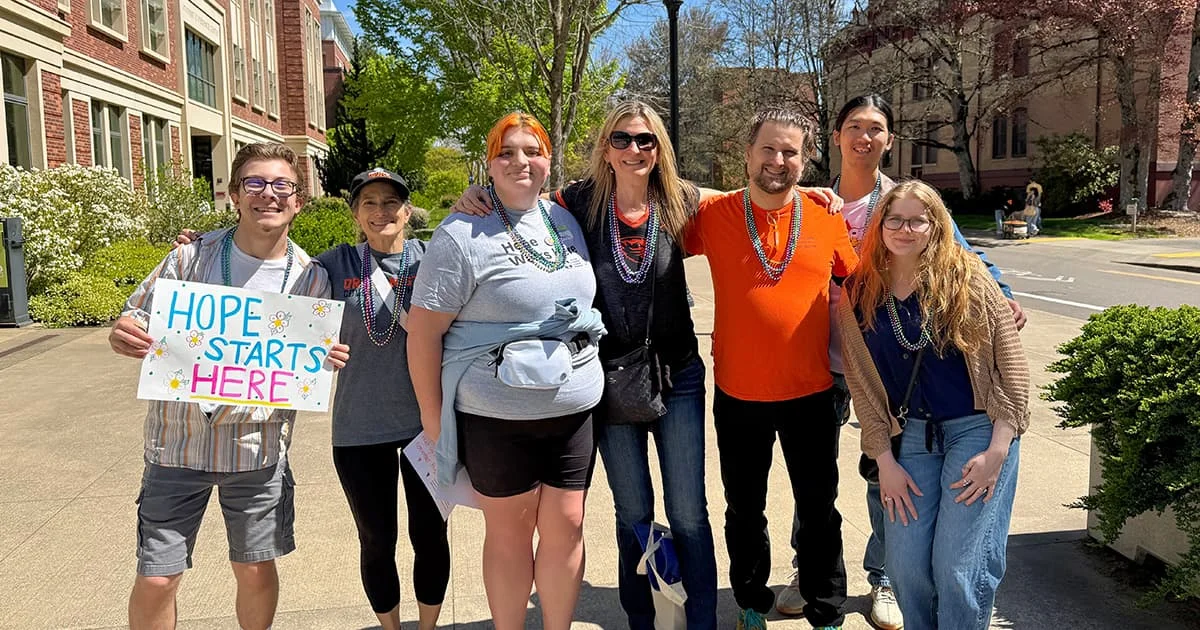 Participants at a community event pose on a sunny campus. A central sign reads 'HOPE STARTS HERE,' emphasizing a message of support and unity.