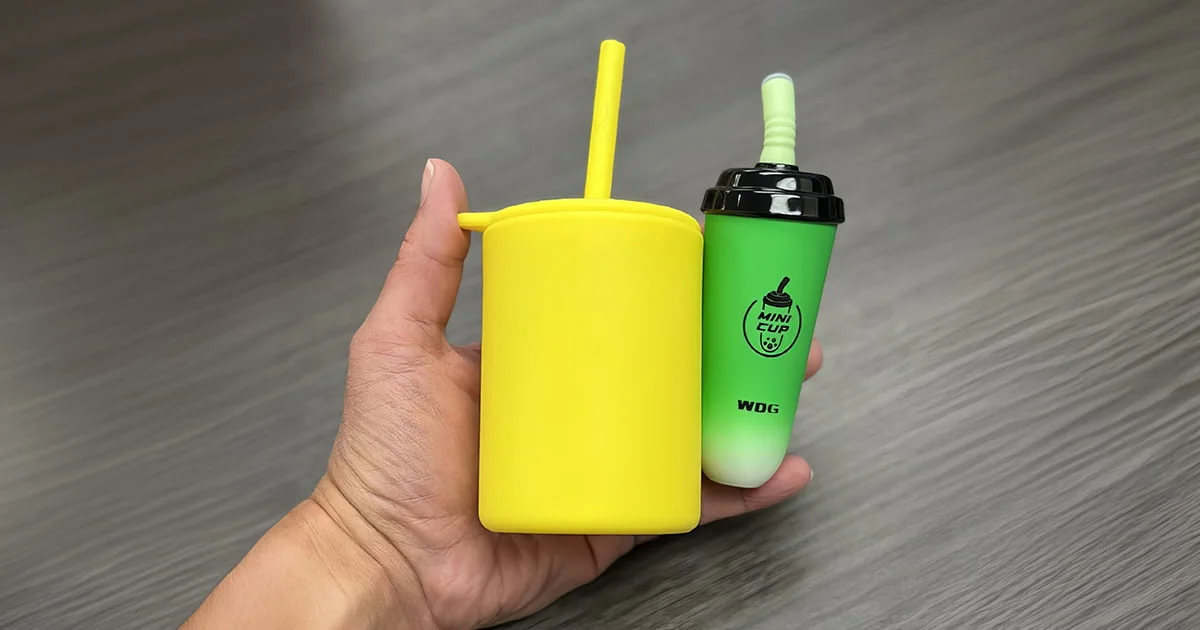A hand holds a yellow children's sippy cup beside a green disposable vape device designed to resemble a mini bubble tea.