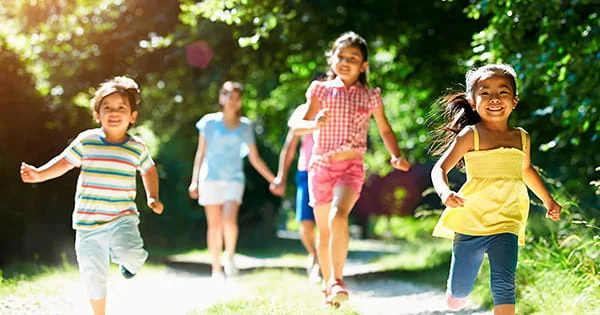 Smiling children running along a sun-dappled outdoor path surrounded by lush green trees.