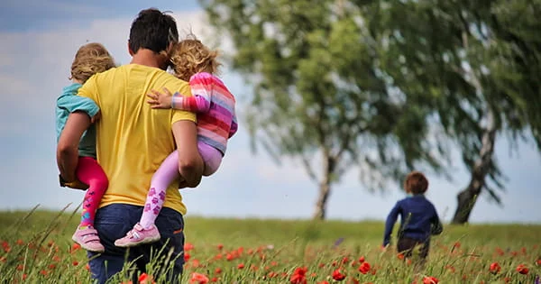 Man carrying two small children in a field of red poppies, with a third child running ahead towards tall trees.
