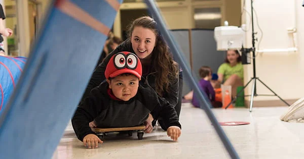 A teacher smiles and assists a child with a physical activity on a scooter board within an indoor sensory gym.