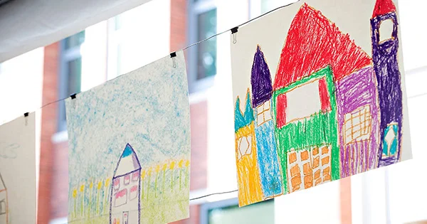 Children's colorful crayon drawings of houses and buildings hanging from a wire with binder clips against a blurred building background.