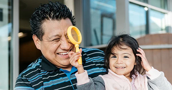 A young child engages in playful discovery, holding a magnifying glass up to a laughing adult's eye.