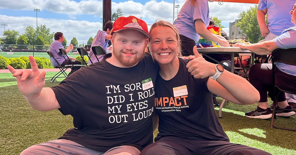 Two participants at an IMPACT inclusive physical activity event smile outdoors. They wear name tags and athletic gear on a bright sports field.