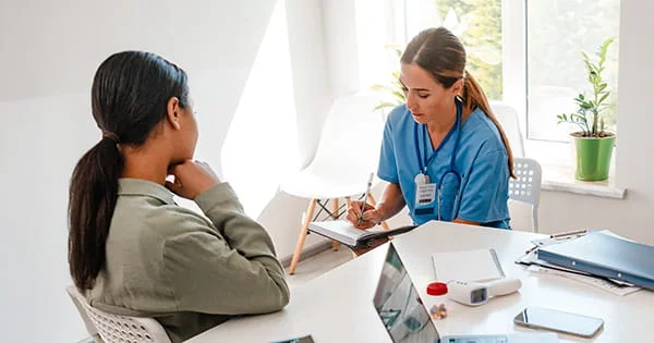 Medical professional in blue scrubs writing notes during a patient consultation in a bright office setting.