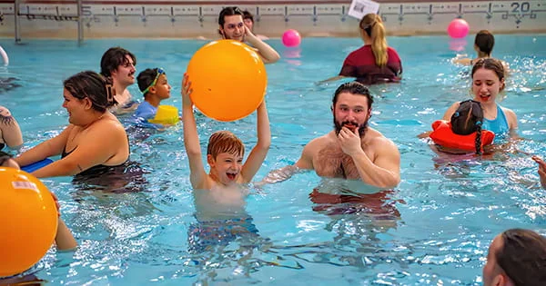 Laughing boy holding a large orange inflatable ball over his head in a crowded indoor swimming pool.