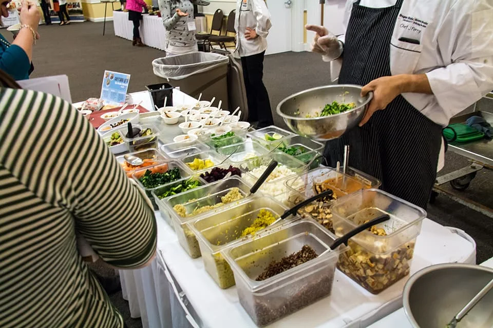 A chef mixes fresh ingredients like leafy greens and quinoa at an event catering table.