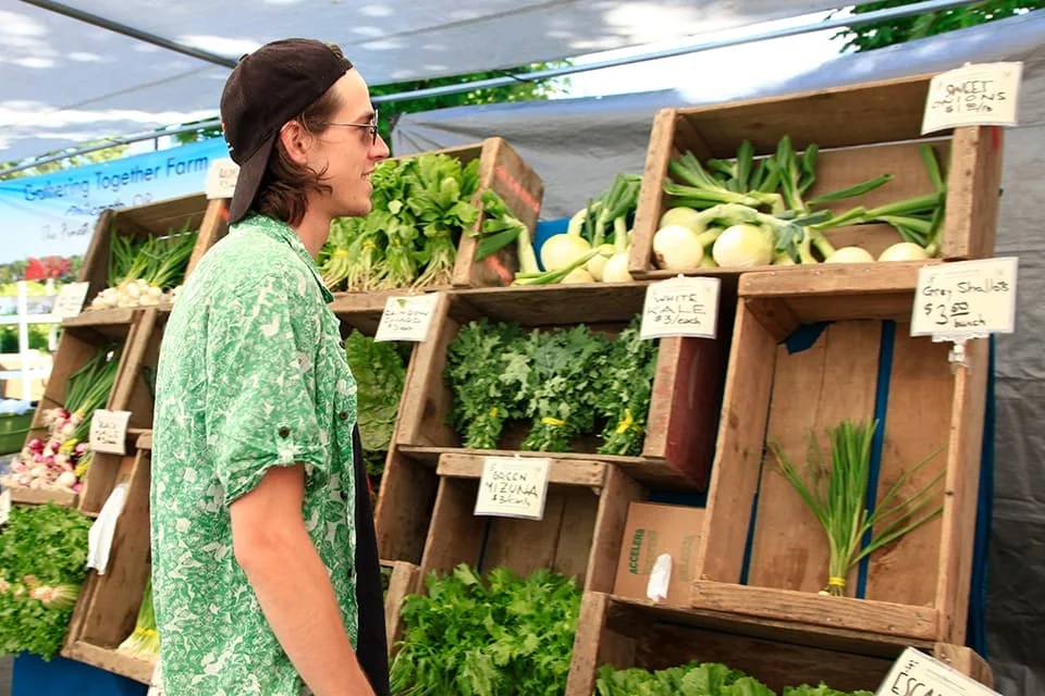 A customer examines produce at a farm stand, with signs for leafy greens and onions visible.