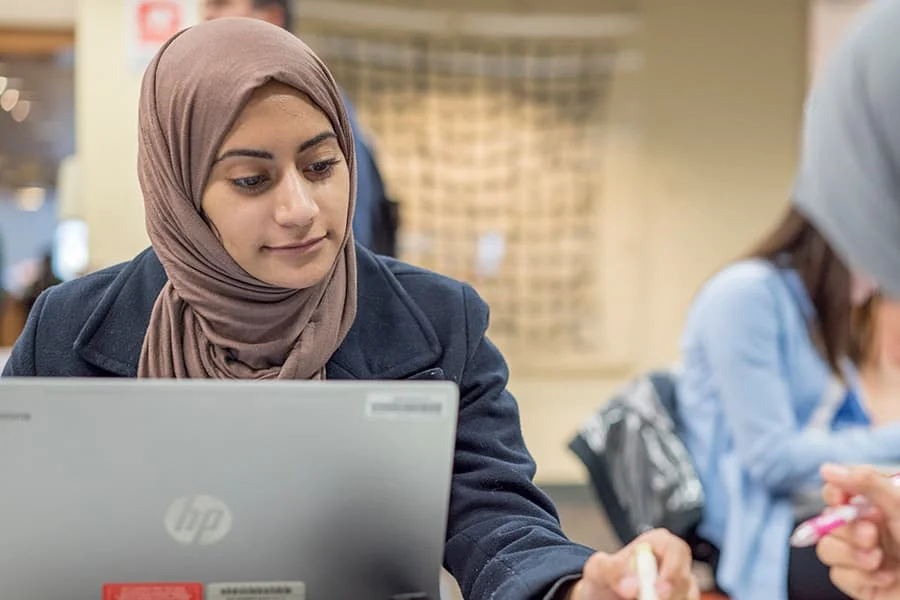 A woman wearing a hijab working on a laptop in an indoor setting.