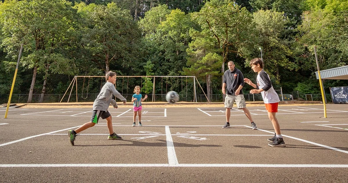 People of different ages engage in active outdoor play, illustrating physical activity and intergenerational interaction in a schoolyard or park setting.