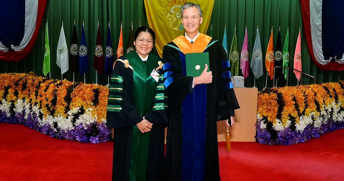 Two people in academic regalia standing on a stage with flags and flowers in the background.