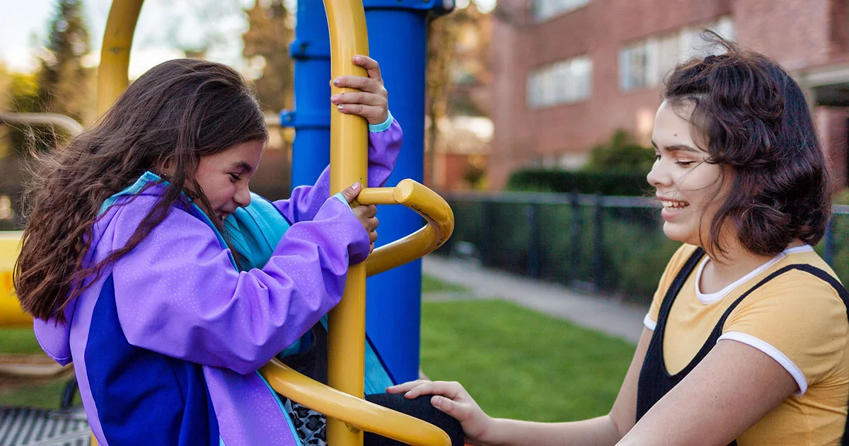 A child in a purple jacket on a playground structure with an adult nearby, looking supportive and caring.