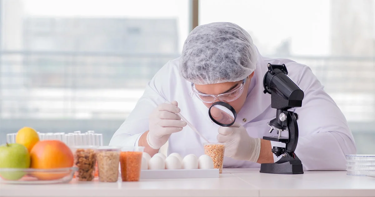 Scientist in white lab coat and protective gear examining something closely with a microscope, with fruits and other items on the table.