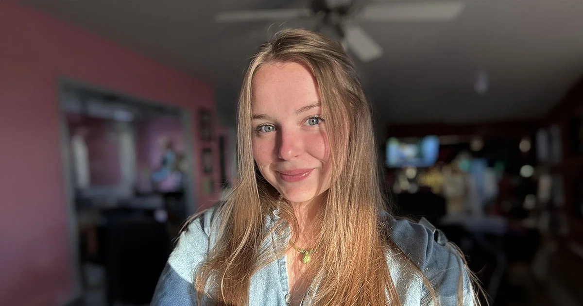 Portrait of a person with long blonde hair wearing a light blue shirt, standing in an indoor setting.