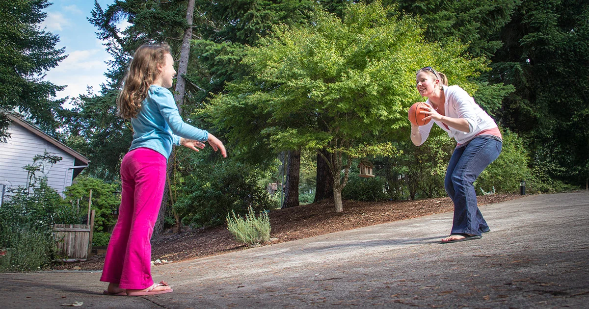 Two people playing basketball together outdoors in a residential area with trees and a house in the background.