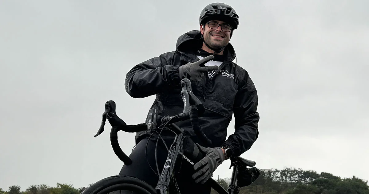 A cyclist wearing a black jacket, helmet, and glasses, sitting on a bicycle against a gray sky background.