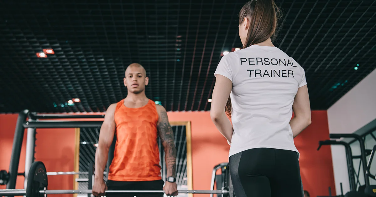 A personal trainer in a white shirt observing a person in an orange tank top lifting weights in a gym.