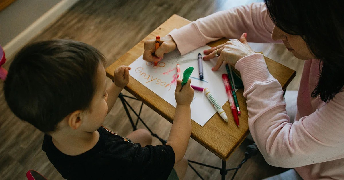 A child and an adult drawing together at a wooden table with colorful markers.