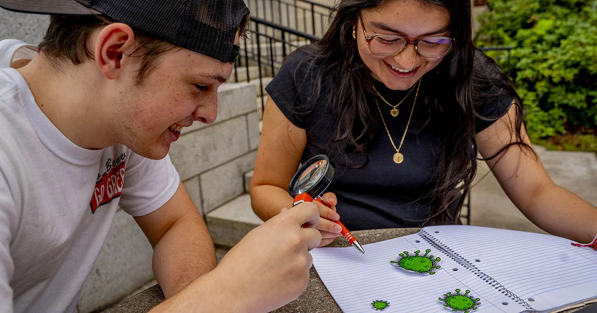 Two students studying together, with a notebook open showing green virus-like illustrations.