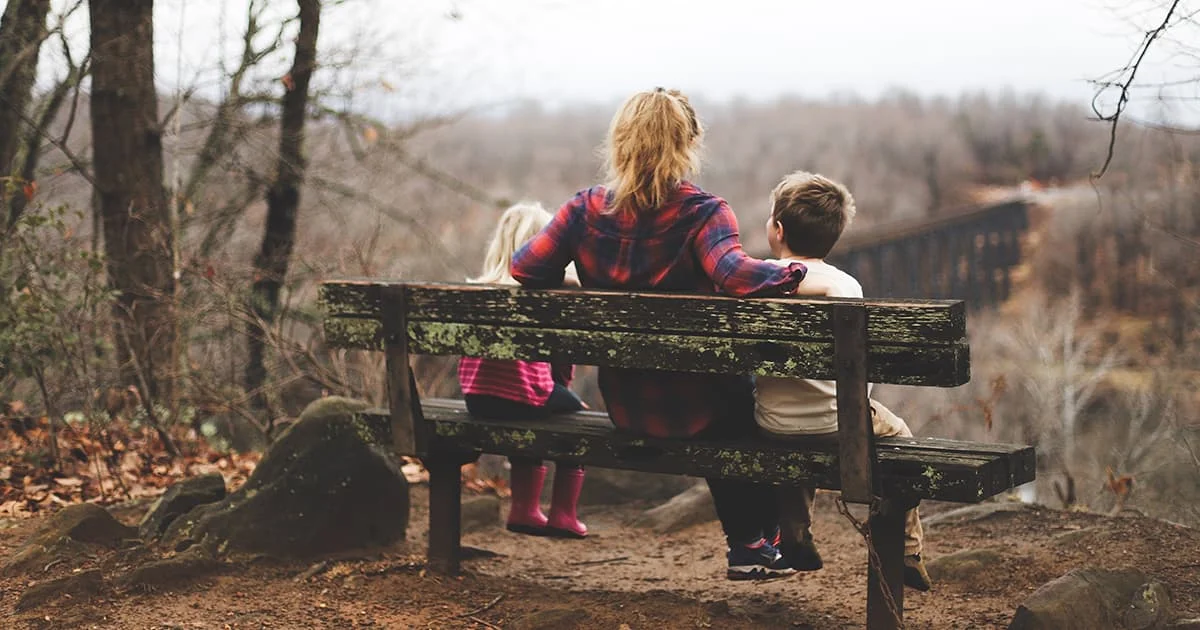 A mother and her two children sit on a bench, looking at a scenic view, symbolizing family bonds, support, and togetherness.