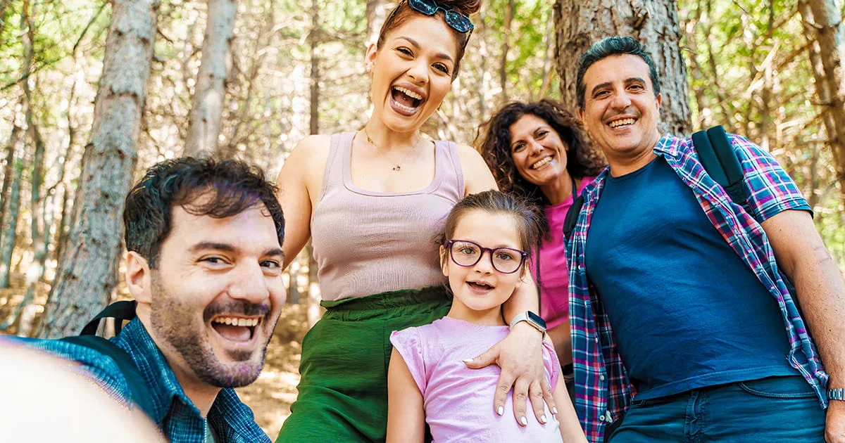 Happy multi-generational family of five taking a selfie while hiking in a sunny, dense forest, emphasizing joy and outdoor activity.