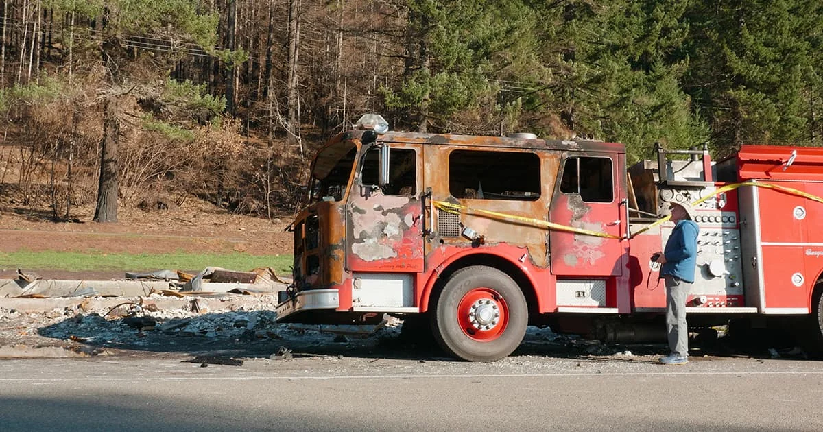 The burned-out, rusted husk of a fire engine sits on the side of a road after a major fire, with a person standing next to it surveying the extensive damage. Burned forest and debris are visible in the background.