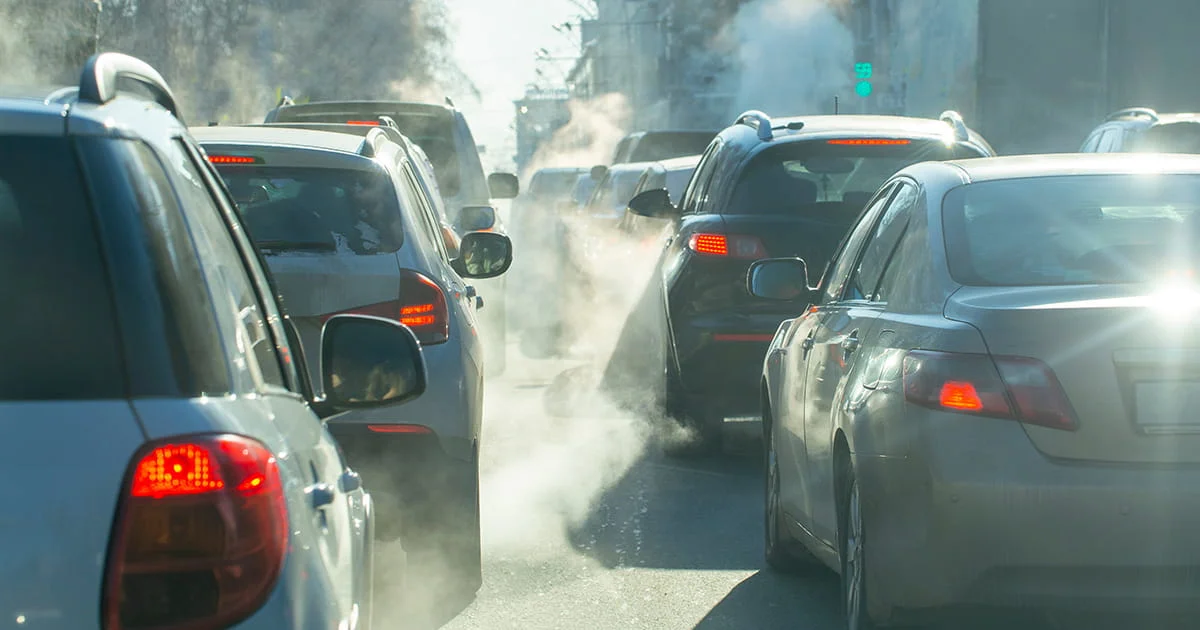 Heavy traffic on a sunny day with vehicle exhaust fumes visible in the air.