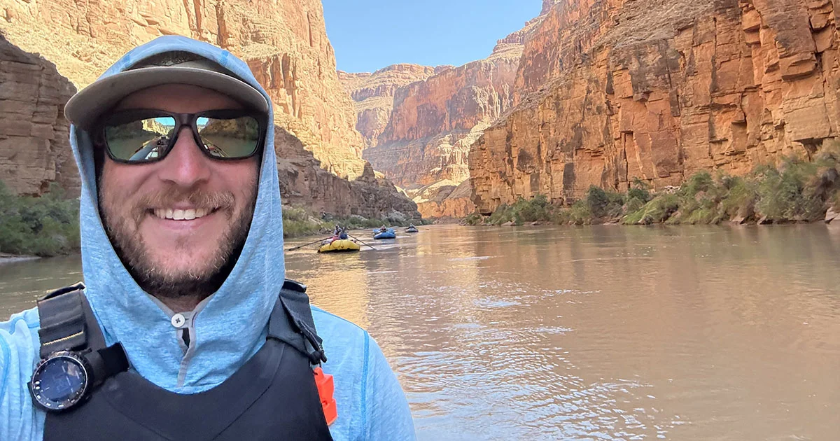 A man wearing a hooded sun shirt, hat and sunglasses smiles while rafting through a canyon on a calm river. Steep orange rock walls rise on both sides and several rafts float in the background.