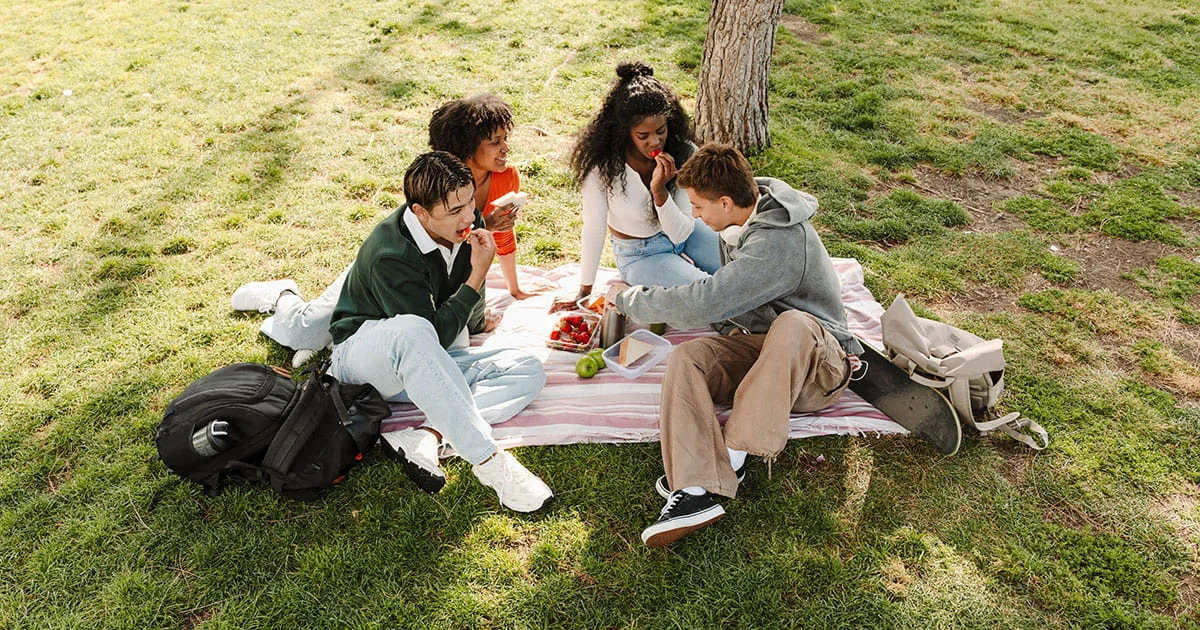 Four friends having a casual outdoor picnic lunch, sharing fresh fruit like strawberries and apples in a grassy, shaded park area.