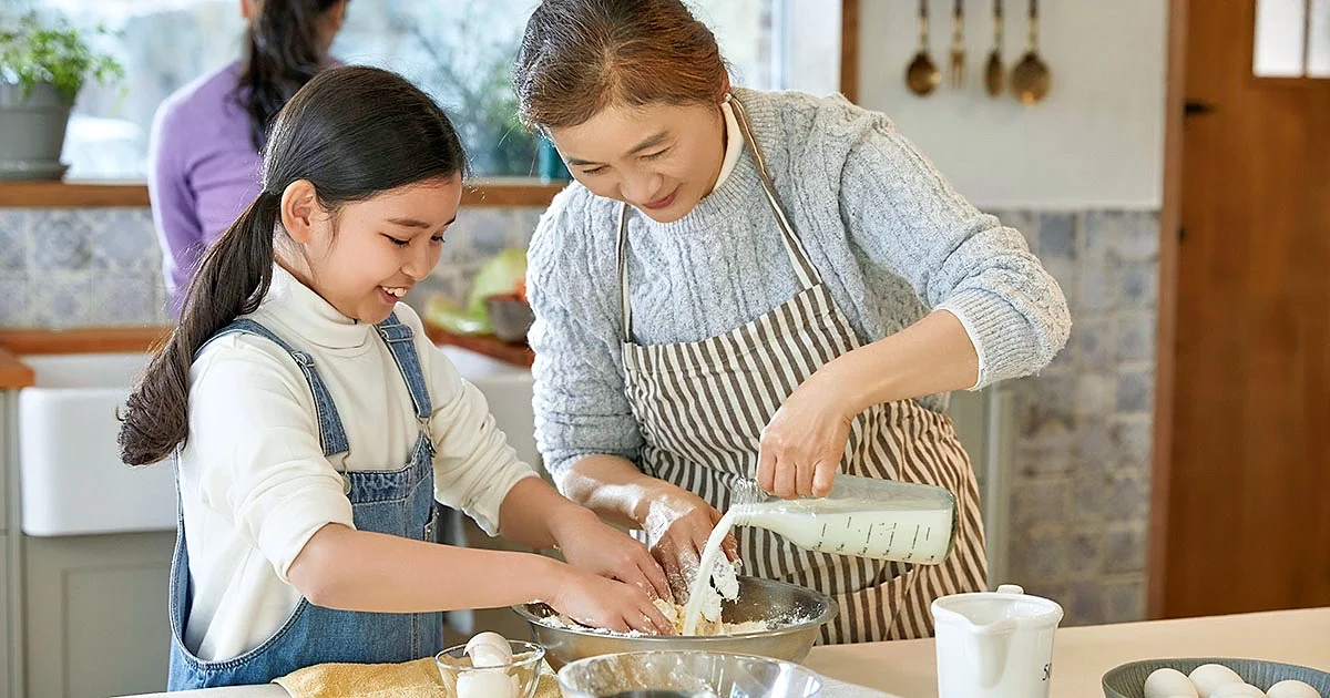 A smiling woman wearing an apron pours milk into a bowl of flour and dough while a young girl next to her helps mix the ingredients by hand.