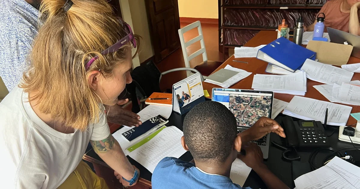 Two people are closely reviewing data and a satellite image on a laptop in a busy office.