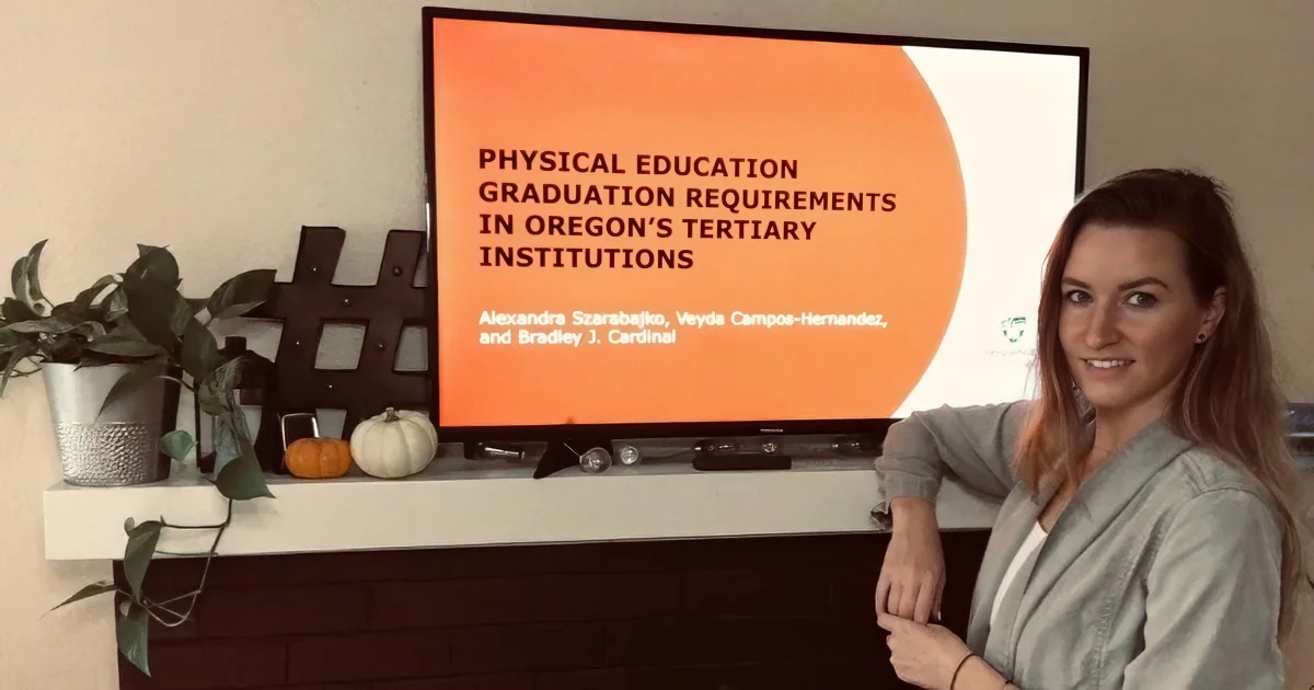 A student presents a slide on a TV screen titled, "Physical Education Graduation Requirements in Oregon's Tertiary Institutions."
