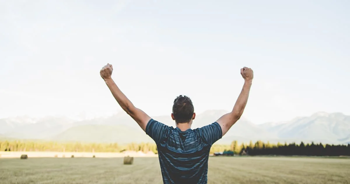 A man standing with his back to the camera in a sunny field, raising his arms in a gesture of success, triumph, or exhilaration.