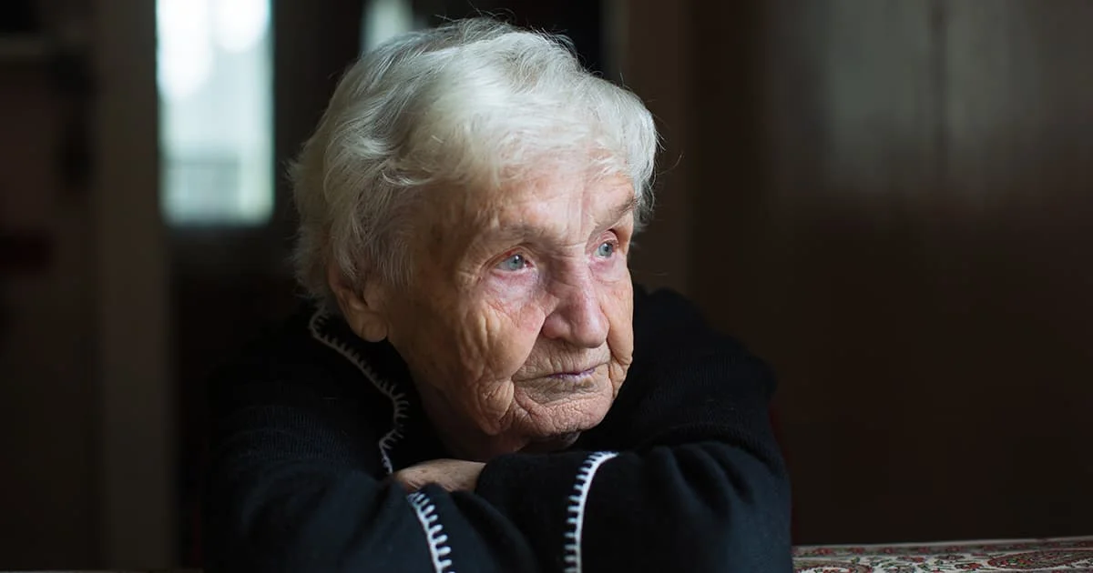 Close-up, somber portrait of an elderly woman with white hair looking away from the camera in a dimly lit room.