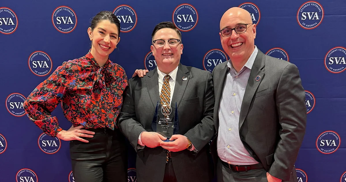 Three smiling individuals pose in front of a blue backdrop patterned with the Student Veterans of America (SVA) logo. The person in the center holds a glass award.