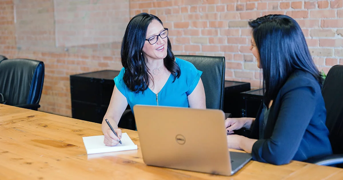 A business meeting or consultation between two colleagues. One woman in a teal shirt smiles while holding a pen, and the other in a dark blazer works at a laptop, illustrating professional teamwork and communication.