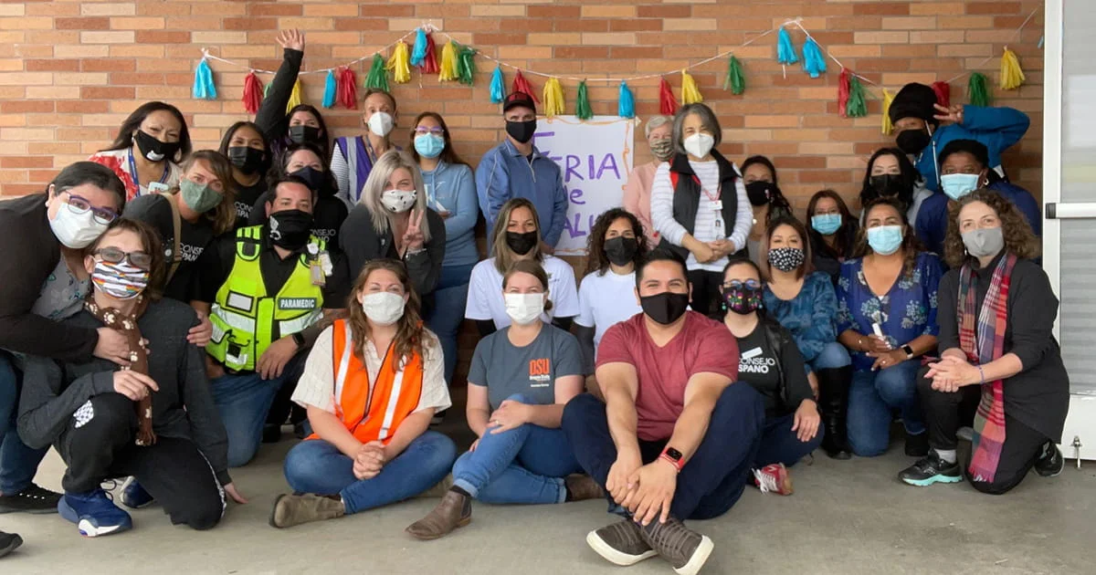 Participants and organizers of a community health fair posing for a group photo. A sign in the background reads "Feria de Salud," symbolizing a collaborative public health outreach event.