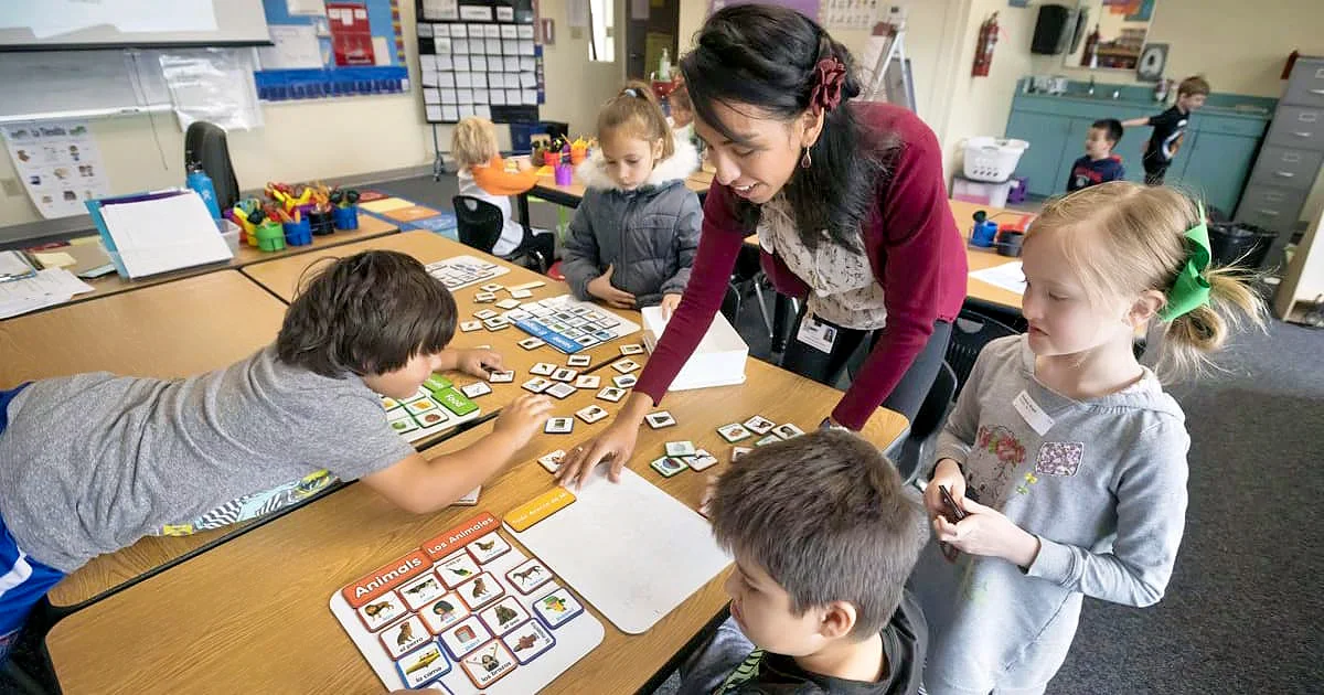 A female teacher in a burgundy cardigan smiles while guiding a group of four young students through a matching game with animal-themed flashcards in a bright classroom.