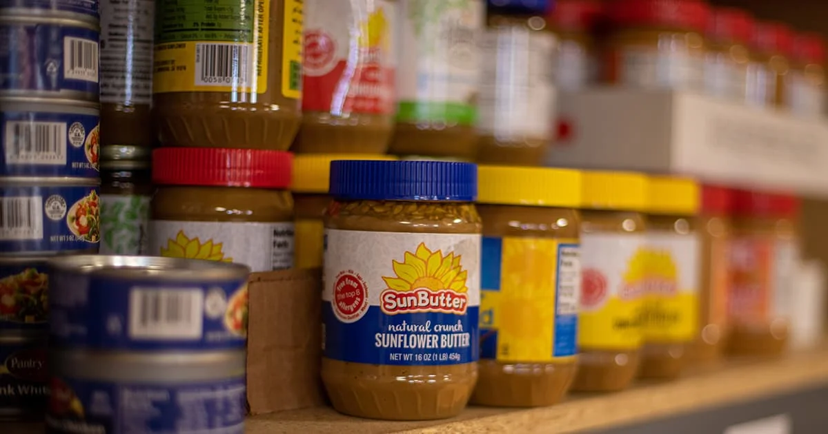 A close-up of a pantry shelf stocked with jars of SunButter Sunflower Butter and stacked cans of food.