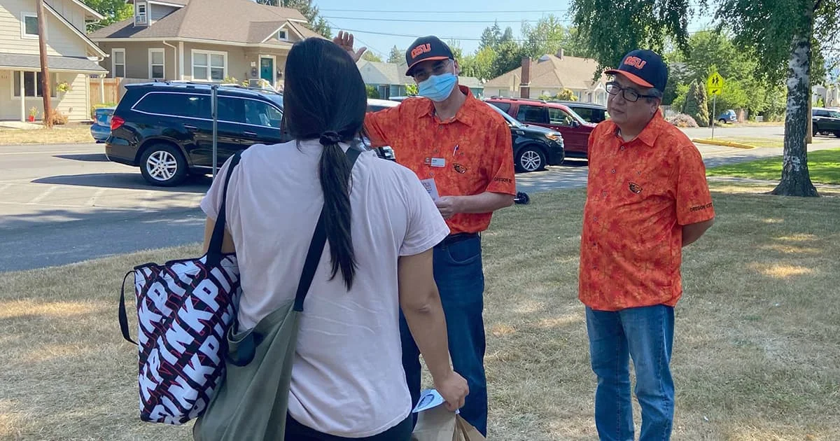 Two men wearing orange Oregon State University (OSU) shirts and face masks talk to a woman outdoors in a residential neighborhood.