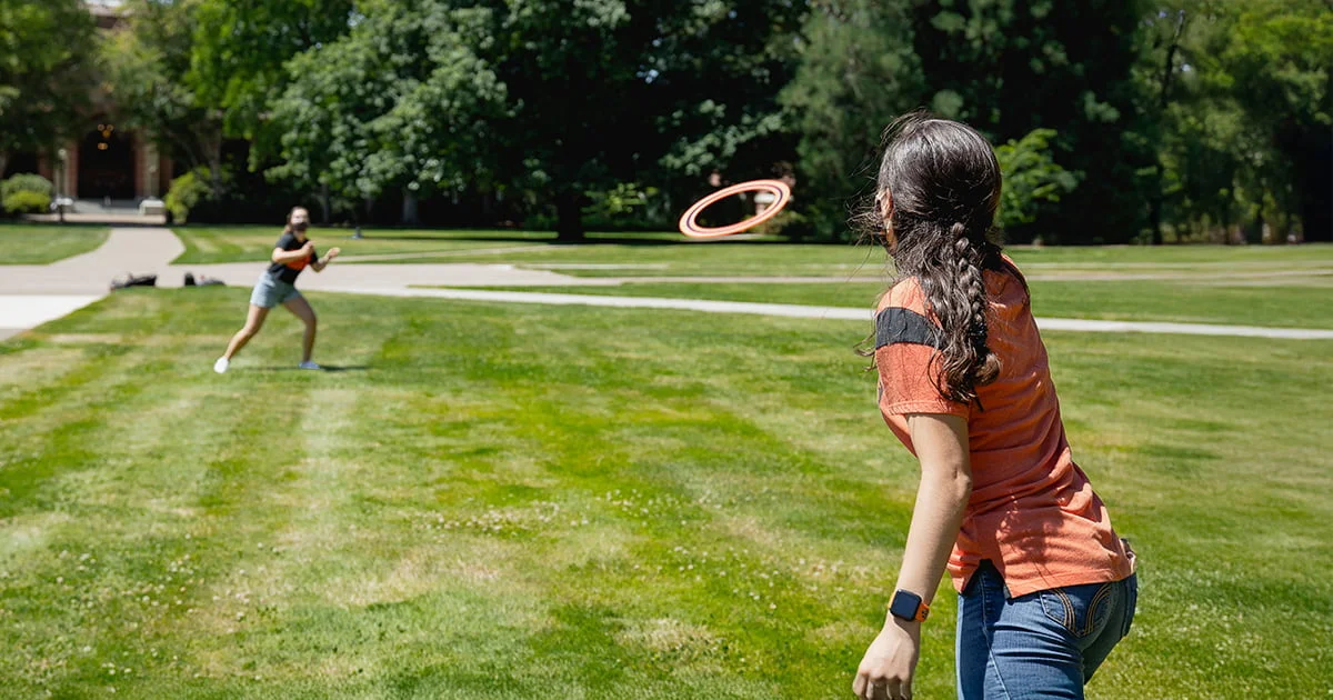 Two students in orange and black athletic shirts play with an orange Aerobie ring on a large green campus lawn.