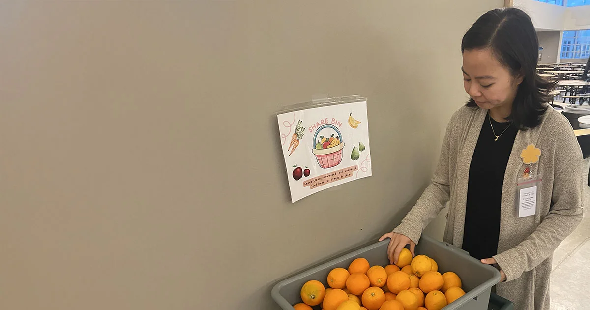 Person wearing a name badge placing oranges into a bin labeled ‘Share Bin,’ with a sign encouraging people to take and share food.